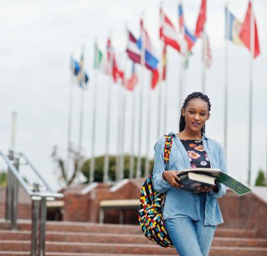 African student female posed with backpack and school items on yard of university, against flags of different countries.